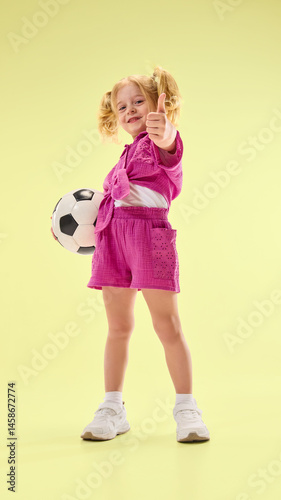Obraz na płótnie Low angle view of happy child girl in pink shorts and shirt, standing with soccer ball and giving thumbs up against yellow background. Concept of childhood, summer vacation, active lifestyle, sport w salonie