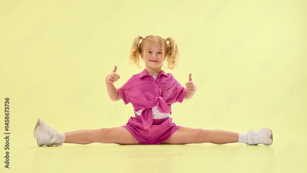 Happy girl child in pink summer clothes and ponytail hairstyle sitting on twine on floor, giving thumbs up and smiling against yellow background. Concept of childhood, summer vacation, fun