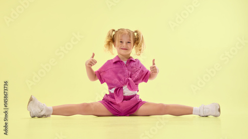 Obraz na płótnie Happy girl child in pink summer clothes and ponytail hairstyle sitting on twine on floor, giving thumbs up and smiling against yellow background. Concept of childhood, summer vacation, fun w salonie