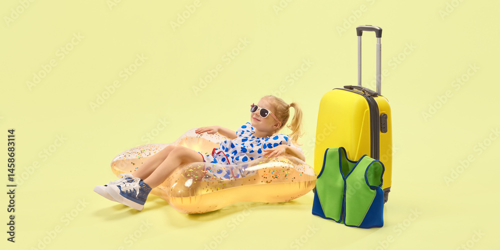 Relaxed girl with ponytail hairstyle, in sunglasses lying on inflatable float beside suitcase joy against yellow background. Concept of childhood, summer vacation, travelling, trip