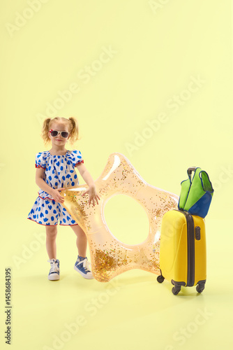 Girl in dress and sunglasses poses with suitcase and float smiling confidently against yellow background. Concept of childhood, summer vacation, travelling, trip