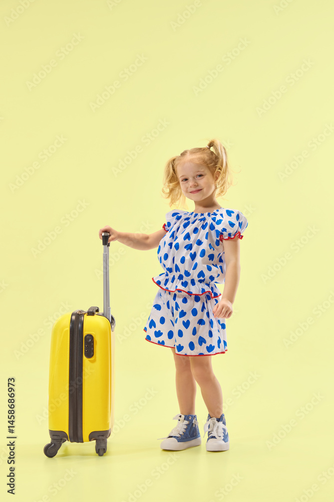 Charming smiling girl standing with suitcase in yellow studio background, wearing dress, ready for adventure. Concept of childhood, summer vacation, travel, fashion, and self-expression.