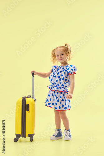 Obraz na płótnie Charming smiling girl standing with suitcase in yellow studio background, wearing dress, ready for adventure. Concept of childhood, summer vacation, travel, fashion, and self-expression. w salonie