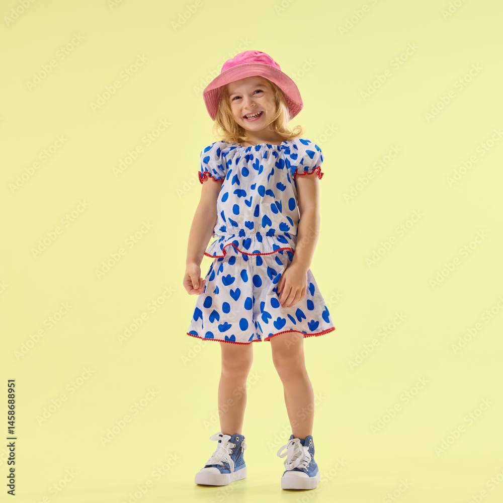 Little girl in cute summer dress, smiling, posing against yellow studio background, showing cheerful mood. Concept of childhood, summer, vacation, play, activity, fun.