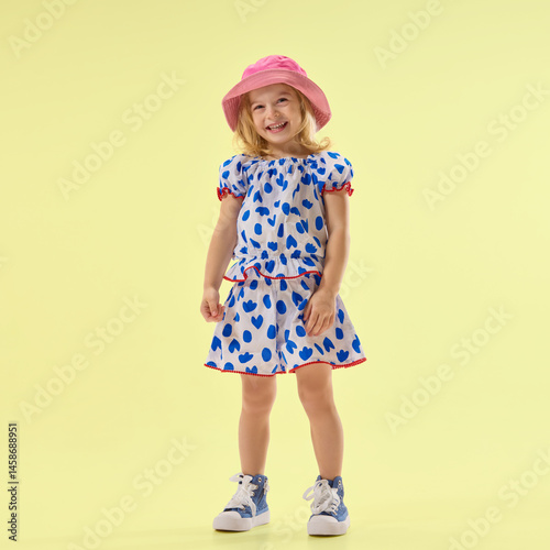 Little girl in cute summer dress, smiling, posing against yellow studio background, showing cheerful mood. Concept of childhood, summer, vacation, play, activity, fun.