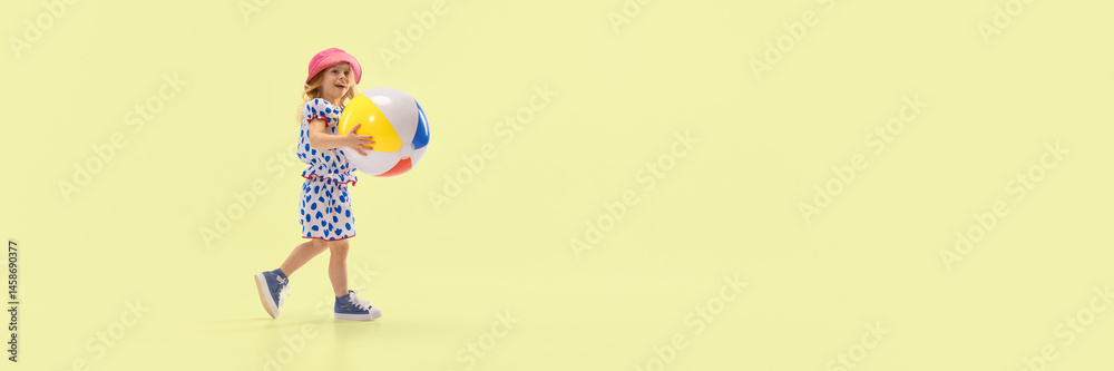 Smiling child walking with big colorful beach ball, enjoying summer play, dressed in polka dot dress and hat against yellow studio background. Concept of childhood, summer, vacation, play, activity
