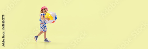 Obraz na płótnie Smiling child walking with big colorful beach ball, enjoying summer play, dressed in polka dot dress and hat against yellow studio background. Concept of childhood, summer, vacation, play, activity w salonie