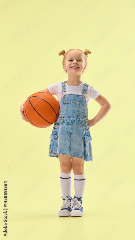 Confident girl smiling, holding basketball on hip, wearing denim overall dress and standing proud, showing positive expression on yellow background. Concept of childhood, summer, vacation, play