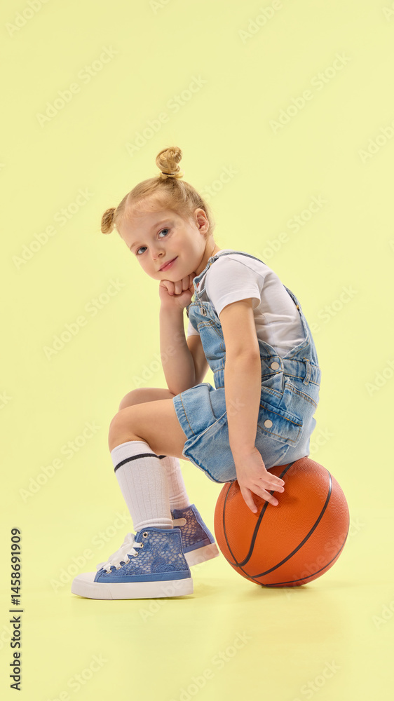 Stylish child sits on basketball, resting chin on hand with thoughtful pose, wearing denim outfit and striped socks in yellow studio background. Concept of childhood, summer, vacation, play, fun.