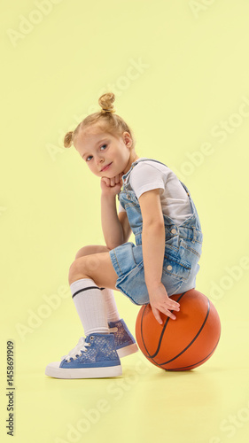 Obraz na płótnie Stylish child sits on basketball, resting chin on hand with thoughtful pose, wearing denim outfit and striped socks in yellow studio background. Concept of childhood, summer, vacation, play, fun. w salonie