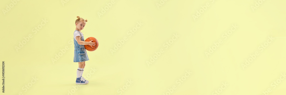 Cute little girl in denim overalls holding basketball, standing in studio with calm expression, ready for game on yellow background. Concept of childhood, summer, vacation, play, activity, fun.