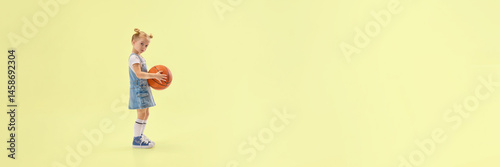 Obraz na płótnie Cute little girl in denim overalls holding basketball, standing in studio with calm expression, ready for game on yellow background. Concept of childhood, summer, vacation, play, activity, fun. w salonie