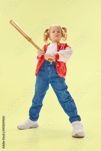 Low angle view of serious girl in red varsity jacket holds bat in action pose against yellow background. Concept of childhood, sport, activity, game, fun, leisure