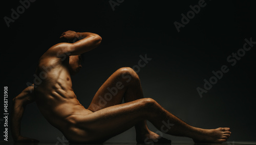 Fotografie A nude male figure sits on the floor with dramatic low directional lighting revealing his muscular form
