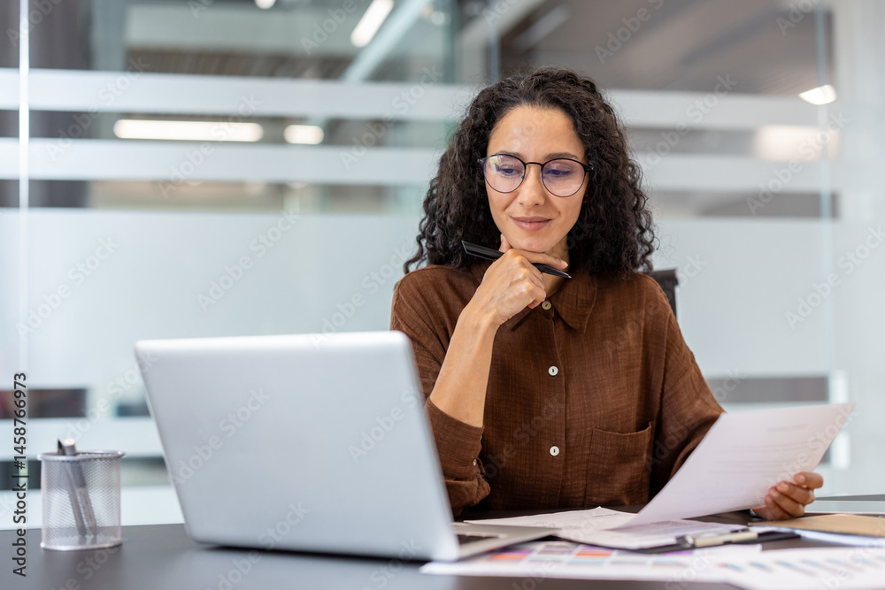 A woman with glasses, working in an office setting, thoughtfully examines documents while using a laptop.