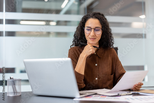 Obraz na płótnie A woman with glasses, working in an office setting, thoughtfully examines documents while using a laptop. w salonie