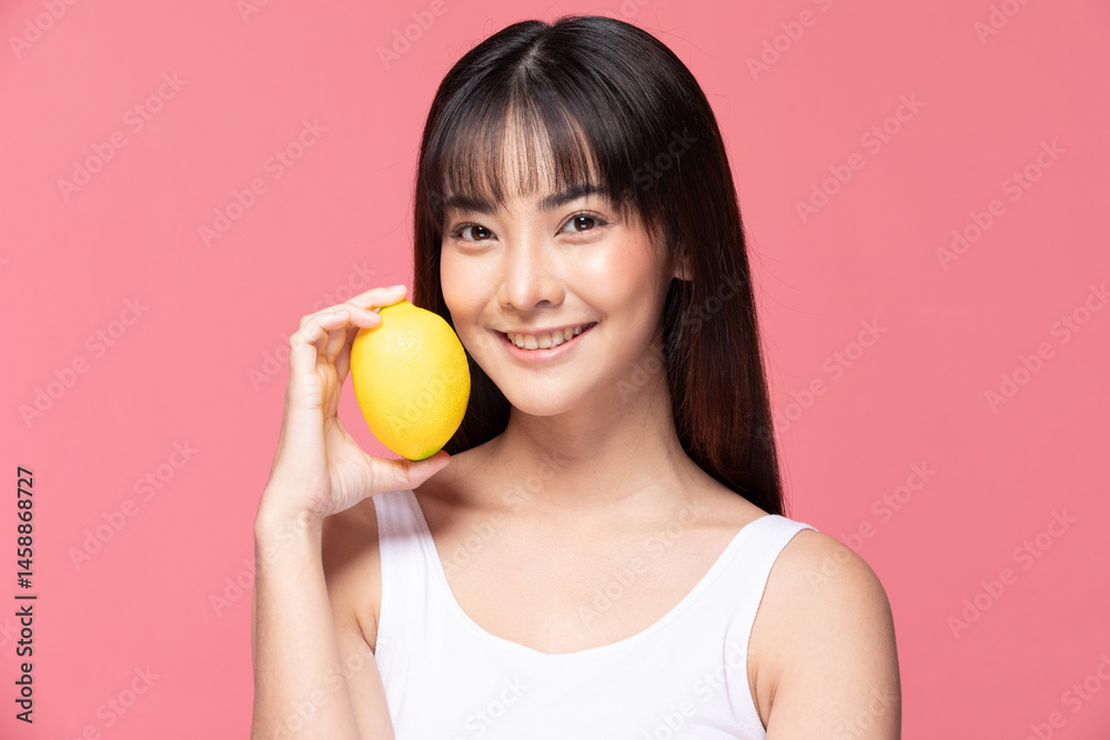 Asian cheerful young woman holding yellow lemon against pink background, wearing white tank top and smiling brightly Asian cheerful young woman holding yellow lemon against pink background, wearing white tank top and smiling brightly