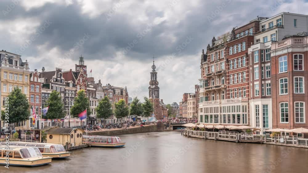 Panoramic timelapse hyperlapse of Amsterdam city center featuring Amstel River, waterfront and Munttoren tower. Boats float back and forth under cloudy sky. Scenic urban view from bridge. Netherlands