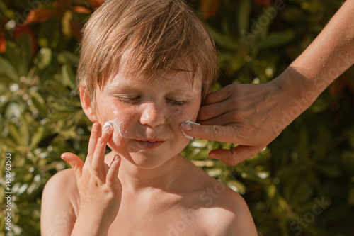 Mother’s hands applying sunscreen to freckled face of young blond boy on a bright sunny day. Summer skincare, sun protection, family care and outdoor parenting concept.
