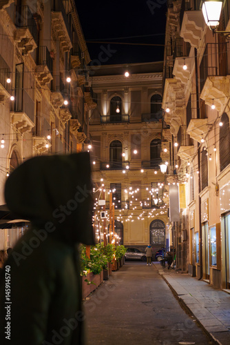 A warmly lit street at night with string lights and elegant buildings, featuring the silhouette of a hooded man walking by.