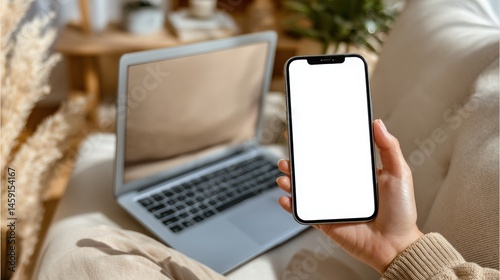 Obraz na płótnie A person holds a smartphone with a blank screen while sitting on a couch near an open laptop in a cozy home setting. w salonie