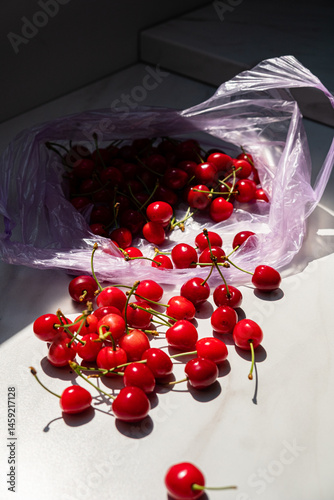 Fototapeta Naklejka Na Ścianę i Meble -  Juicy red cherries tumble out of a plastic bag, brightening up a sunlit kitchen space