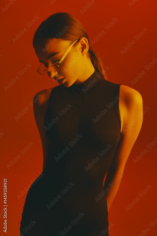 Portrait of young attractive woman with ponytail, wearing transparent trendy glasses and sleeveless bodysuit, posing against red background in neon light. Concept of fashion, style, youth culture