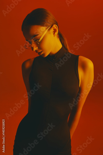 Obraz na płótnie Portrait of young attractive woman with ponytail, wearing transparent trendy glasses and sleeveless bodysuit, posing against red background in neon light. Concept of fashion, style, youth culture w salonie