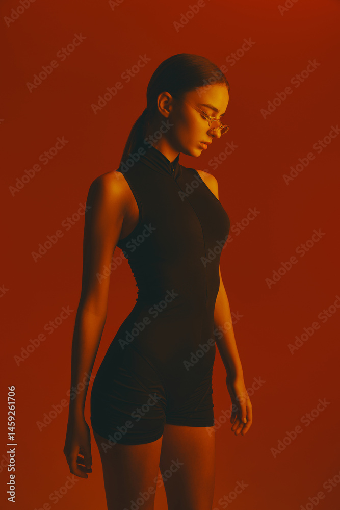 Portrait of beautiful and stylish young woman with slim body, wearing black bodysuit and transparent glasses against red studio background in neon light. Concept of fashion, style, youth culture