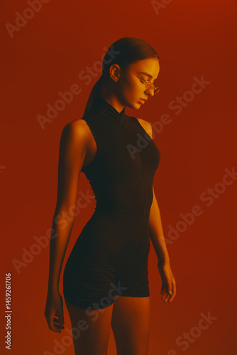 Obraz na płótnie Portrait of beautiful and stylish young woman with slim body, wearing black bodysuit and transparent glasses against red studio background in neon light. Concept of fashion, style, youth culture w salonie