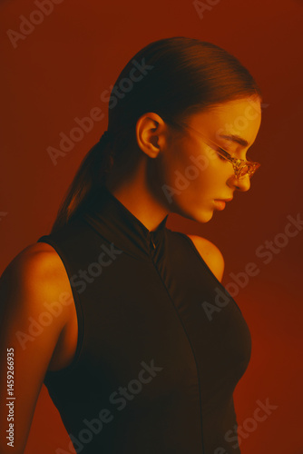 Obraz na płótnie Side view portrait of elegant young woman in trendy glasses and black bodysuit posing against red studio background in neon light. Concept of fashion, style, youth culture, trends w salonie