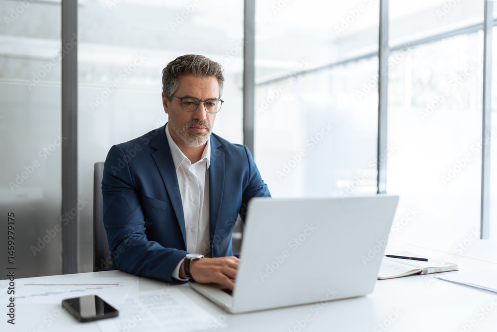 Fototapeta premium Busy latin male executive businessman looking on computer thinking, working on finance data sitting at desk in office. Mature indian business man in eyeglasses, suit using pc laptop for trading work