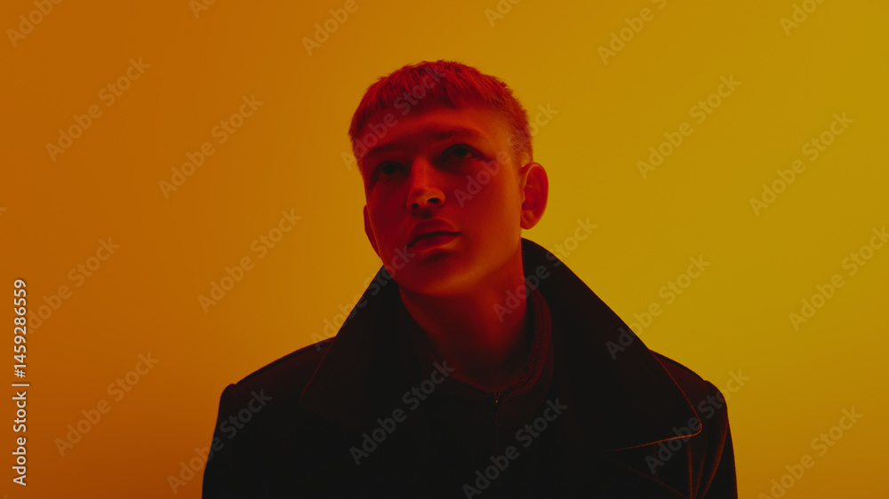 Portrait of stylish young guy in black coat and short hair style, looking upwards, posing against gradient yellow background in neon light. Bold fashion, style, youth culture, self-expression concept