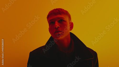 Obraz na płótnie Portrait of stylish young guy in black coat and short hair style, looking upwards, posing against gradient yellow background in neon light. Bold fashion, style, youth culture, self-expression concept w salonie