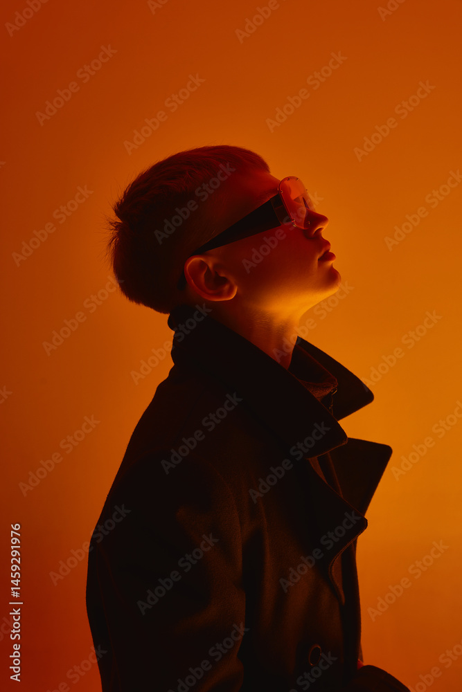 Side view portrait of young guy with short hair, wearing black coat and futuristic big sunglasses, looking upwards against orange studio background. Bold fashion, style, youth culture, self-expression