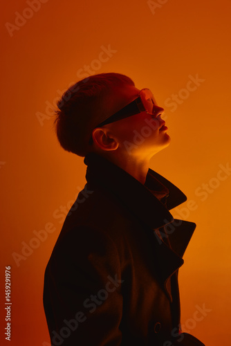 Obraz na płótnie Side view portrait of young guy with short hair, wearing black coat and futuristic big sunglasses, looking upwards against orange studio background. Bold fashion, style, youth culture, self-expression w salonie