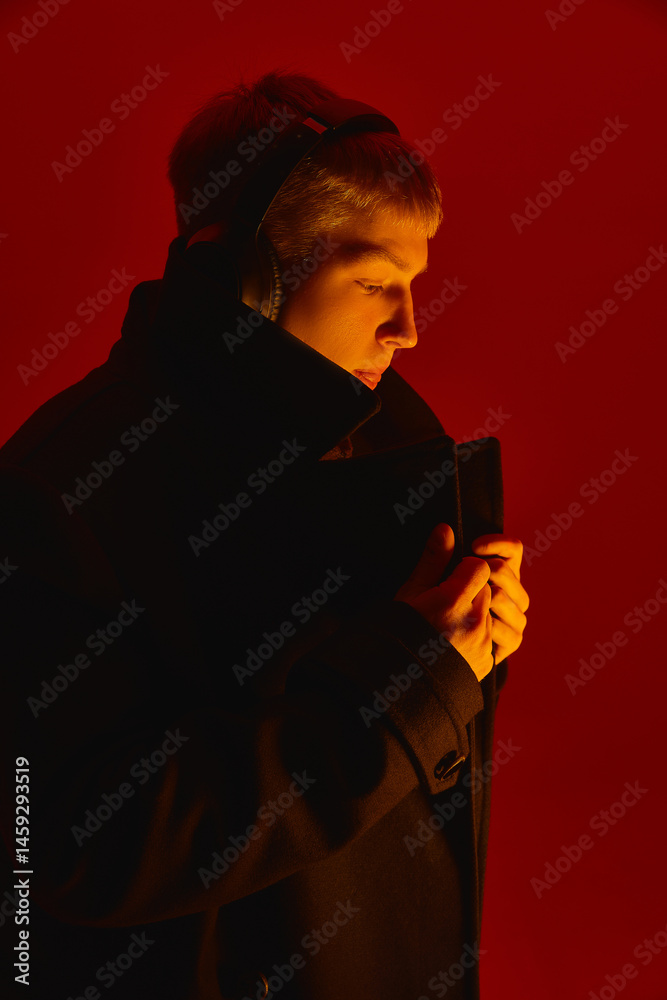 Stylish young man in headphones and black coat looks aside with serious expression under deep red studio lighting setup. Concept of bold fashion, style, youth culture, self-expression