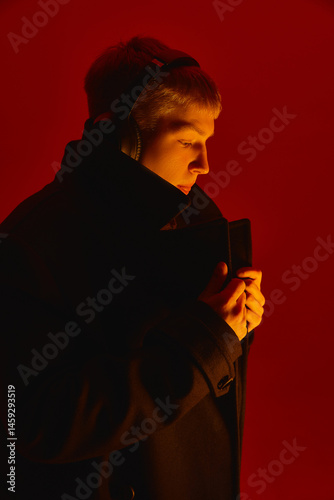 Obraz na płótnie Stylish young man in headphones and black coat looks aside with serious expression under deep red studio lighting setup. Concept of bold fashion, style, youth culture, self-expression w salonie