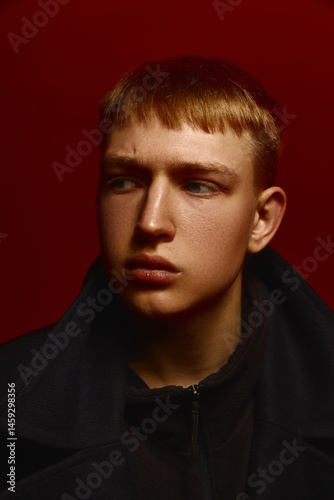 Stylish young man in black coat looks aside with serious expression under deep red studio lighting setup. Concept of bold fashion, style, youth culture, self-expression
