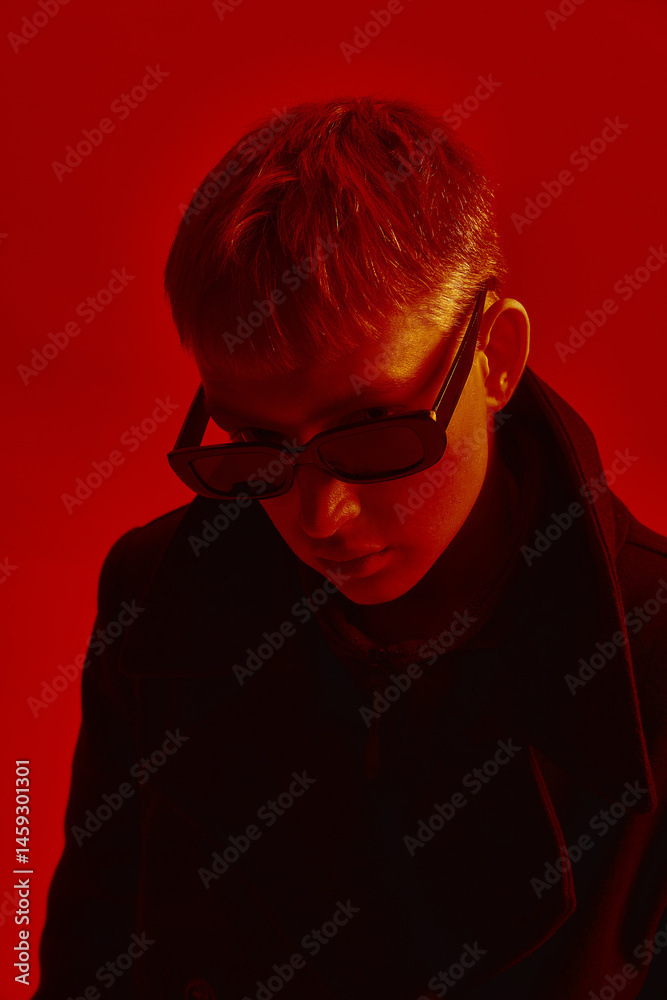 Young man wearing sunglasses and black coat, posing under deep red light against red studio background. Concept of bold fashion, style, youth culture, self-expression