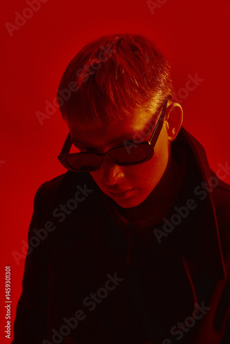 Obraz na płótnie Young man wearing sunglasses and black coat, posing under deep red light against red studio background. Concept of bold fashion, style, youth culture, self-expression w salonie