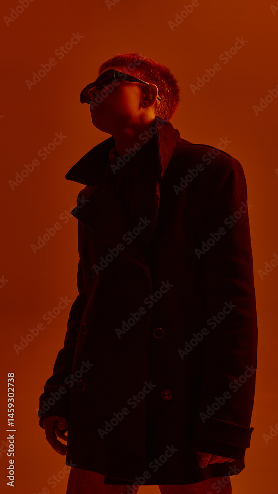 Low angle portrait of young stylish guy in sunglasses and black coat, posing against red studio background in neon light. Concept of bold fashion, style, youth culture, self-expression