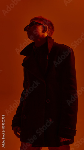 Obraz na płótnie Low angle portrait of young stylish guy in sunglasses and black coat, posing against red studio background in neon light. Concept of bold fashion, style, youth culture, self-expression w salonie