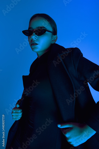 Portrait of confident young woman in sunglasses, black coat and body suit, posing against blue background in neon light. Concept of bold fashion, style, youth culture, self-expression, beauty