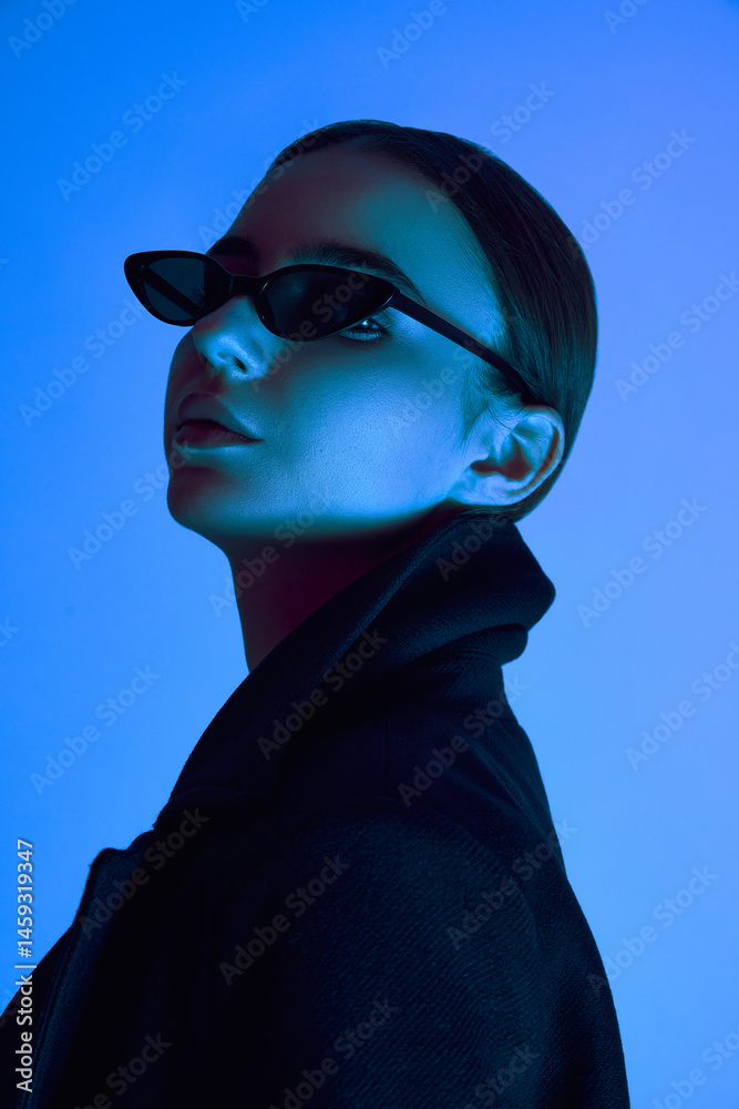 Close-up side portrait of elegant young woman in sunglasses and black coat posing confidently against blue background in neon light. Concept of bold fashion, youth culture, self-expression, beauty