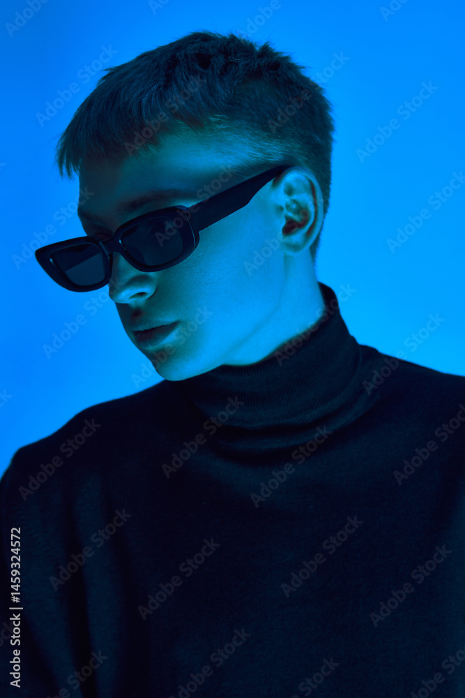 Close-up portrait of stylish young man in sunglasses and black coat posing confidently against blue background in neon light. Concept of bold fashion, youth culture, self-expression, beauty