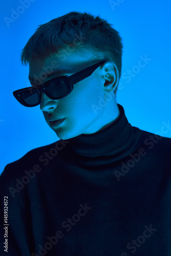 Obraz na płótnie Close-up portrait of stylish young man in sunglasses and black coat posing confidently against blue background in neon light. Concept of bold fashion, youth culture, self-expression, beauty w salonie