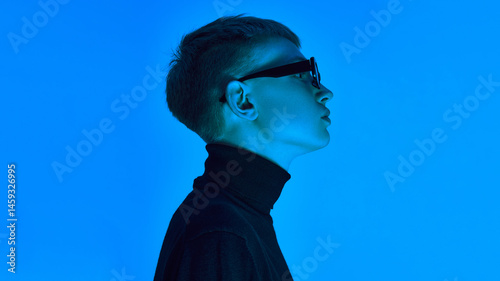Side profile of stylish young man in turtleneck and sunglasses, posing under deep neon blue background ,expresses futuristic elegance. Concept of bold fashion, style, youth culture, self-expression