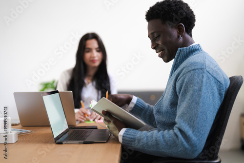 A young man reviews notes in a book at the office. A colleague works on a laptop in the background.