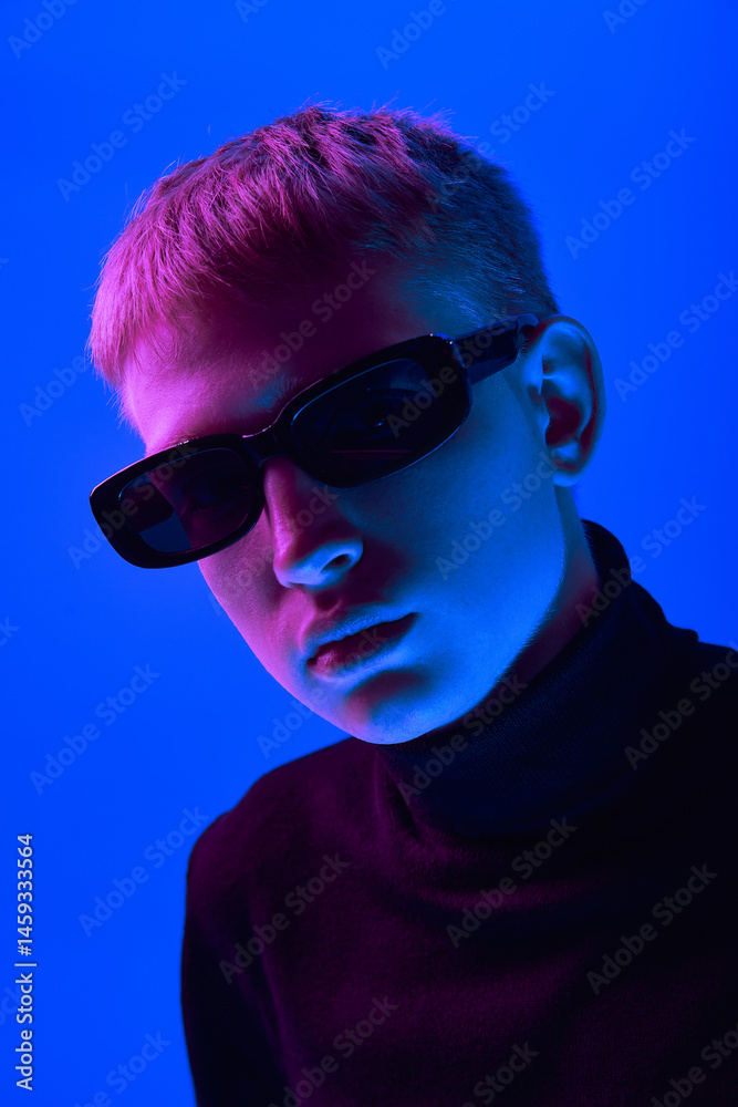 Portrait of stylish young guy in sunglasses and black turtleneck posing against blue background in neon light. Close-up. Concept of bold fashion, style, youth culture, self-expression
